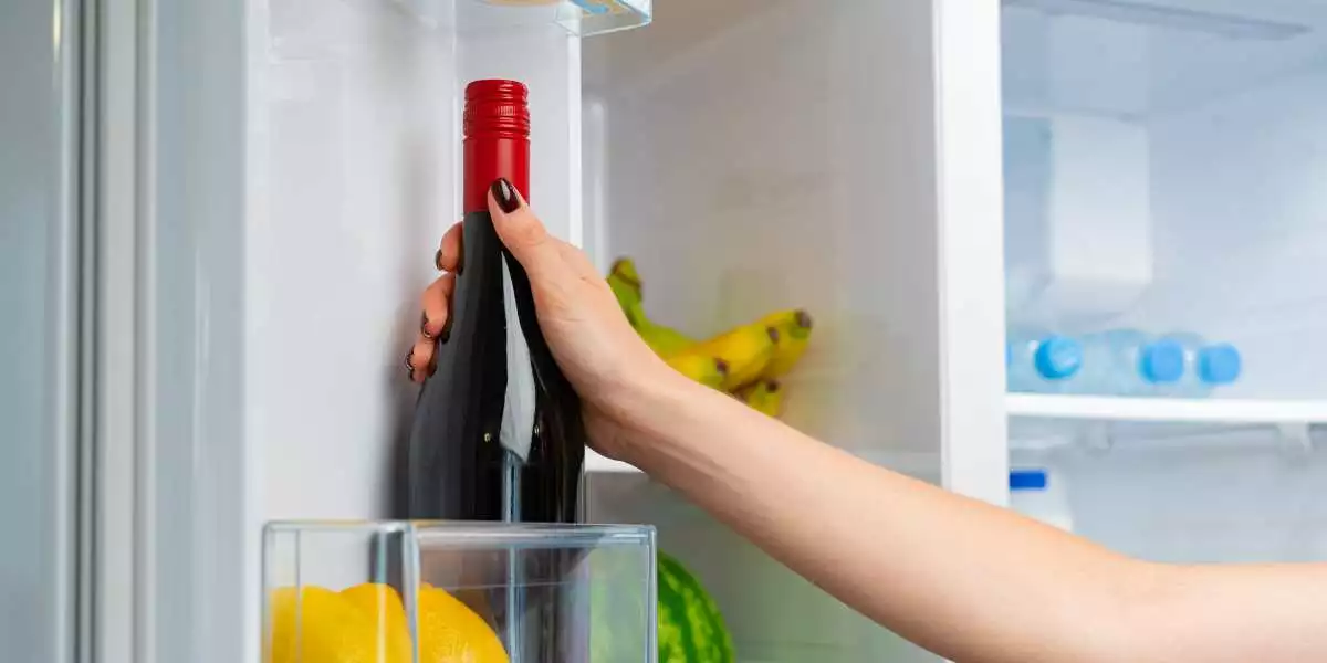 A woman is holding a bottle of wine in an open refrigerator.