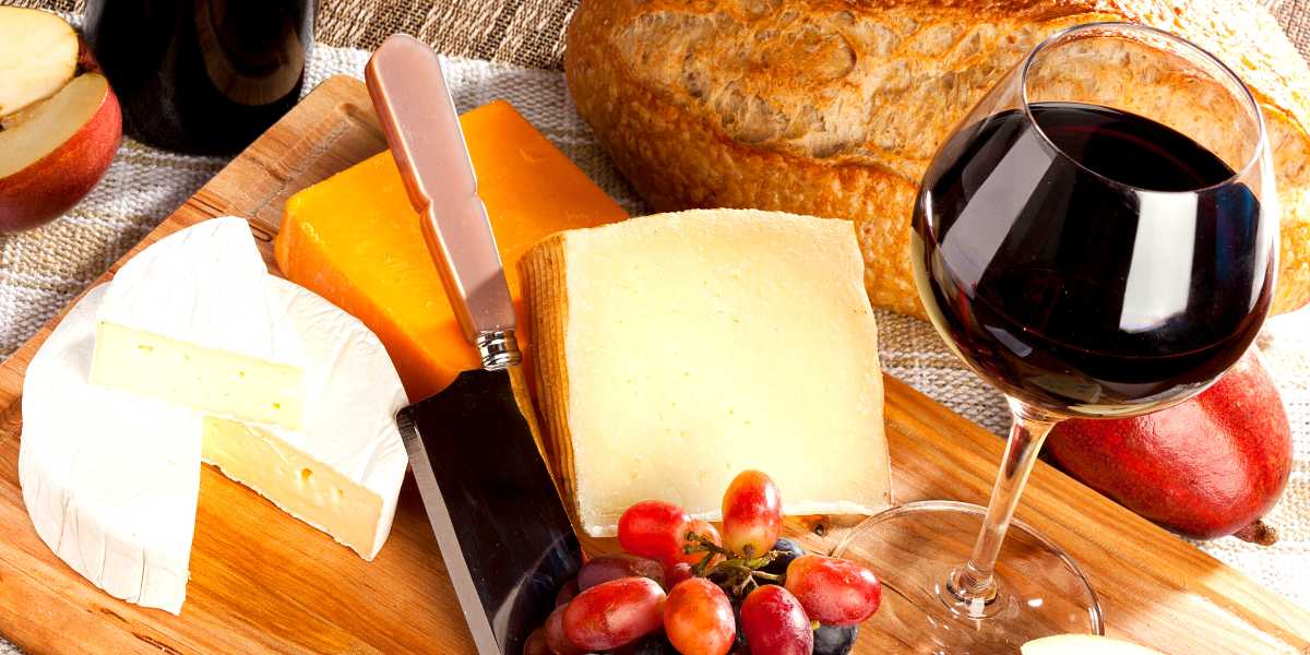 A glass of wine, cheese, fruit and bread on a wooden cutting board.